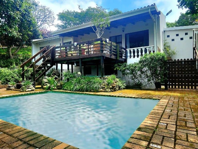 Rear exterior view featuring a swimming pool, raised wooden deck, and brick paved patio area set within a garden.