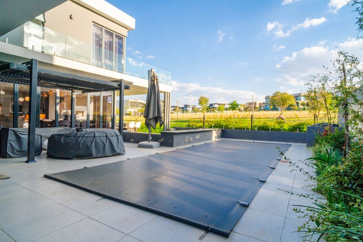 A modern rear patio area featuring a covered pergola, tiled flooring, and a swimming pool with a safety cover, overlooking a landscaped estate.