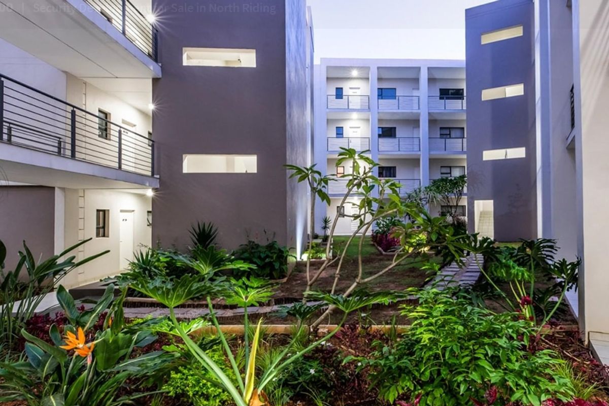 A well-maintained apartment complex courtyard featuring landscaped garden beds, paved pathways, and modern multi-story building facades with balcony access.
