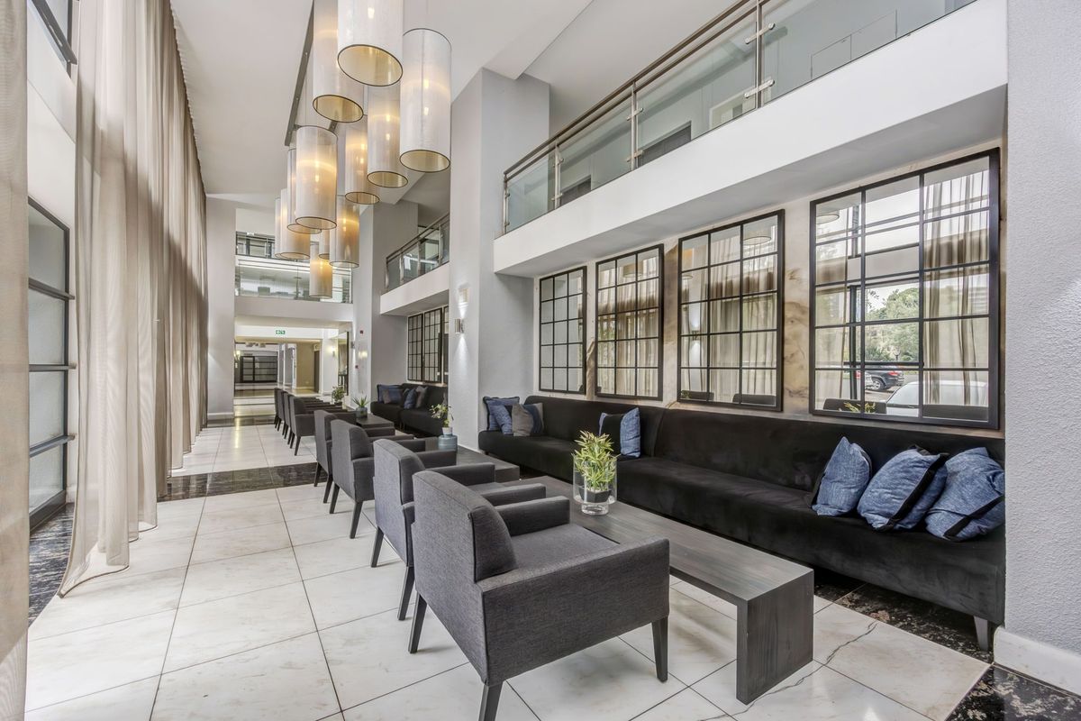 Spacious double-volume living area featuring modern tiled flooring, a glass-railed mezzanine level, and elegant pendant lighting in a contemporary apartment building.