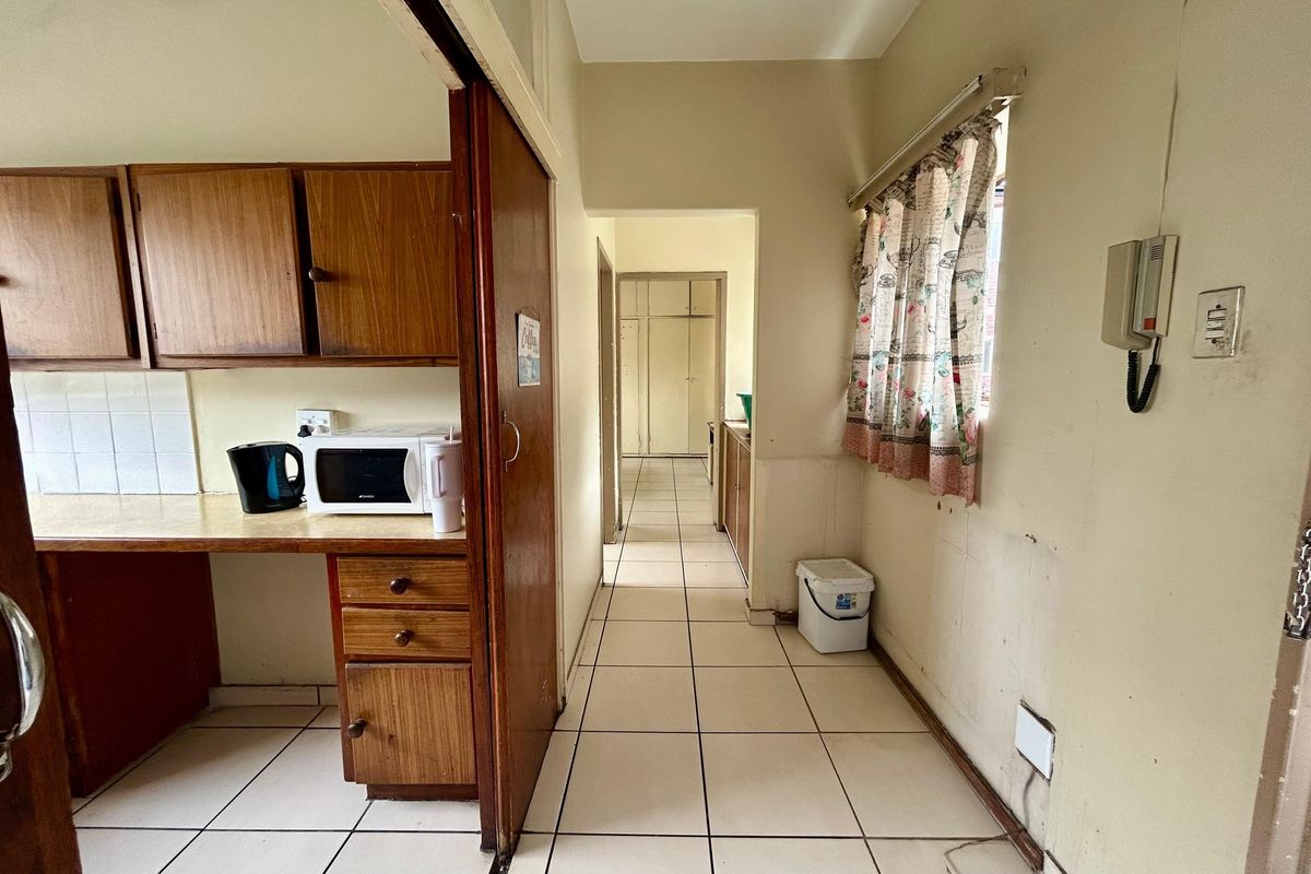 A dated kitchen area featuring wooden cabinetry and tiled flooring, leading into a hallway. The space requires cosmetic updates to the wall finishes.