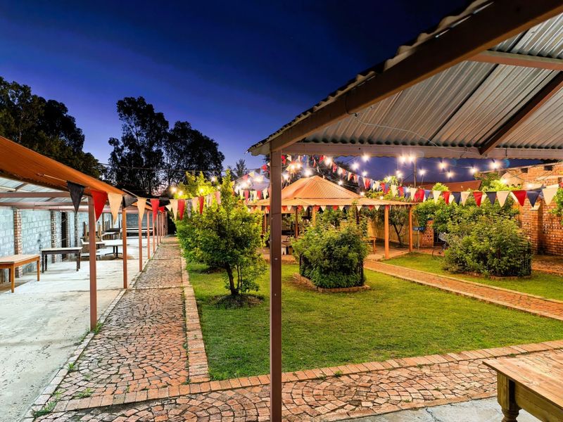 An expansive outdoor entertainment area featuring covered walkways, brick-paved paths, a central gazebo, and established garden landscaping under a twilight sky.