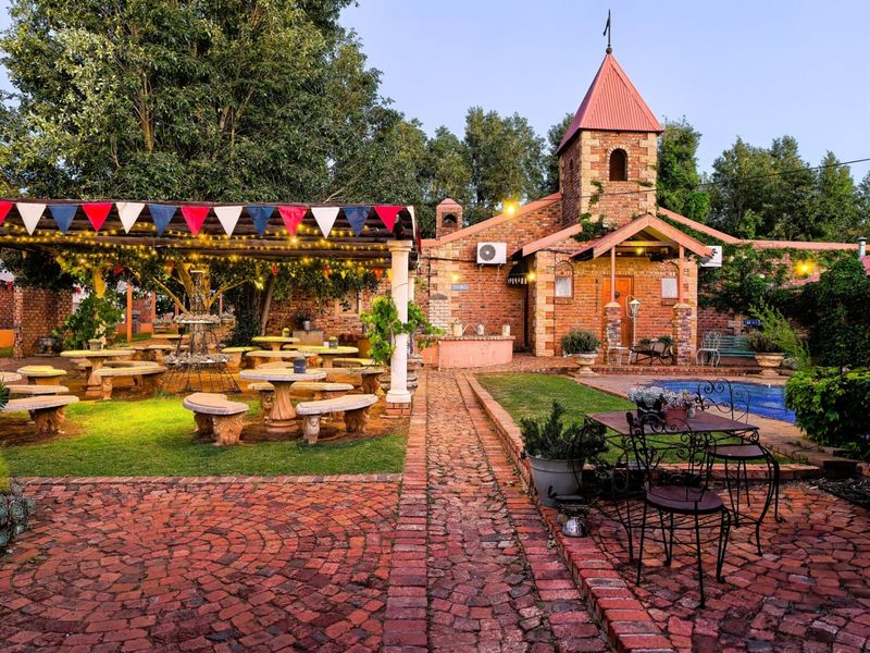 A charming rear garden area featuring brick paving, a covered patio with stone seating, and a unique brick building with a tower and swimming pool.