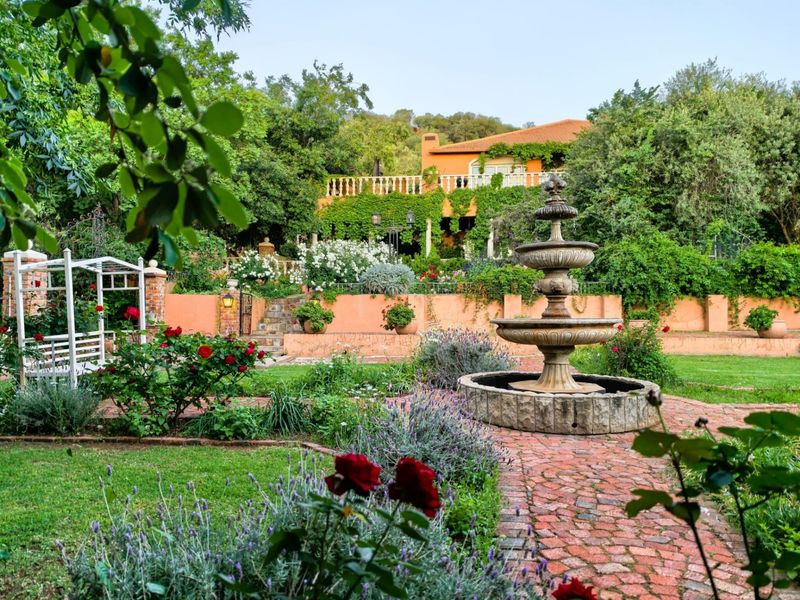 A beautifully landscaped garden featuring a central stone fountain, brick pathways, and a multi-level terrace with stone balustrades and lush greenery.