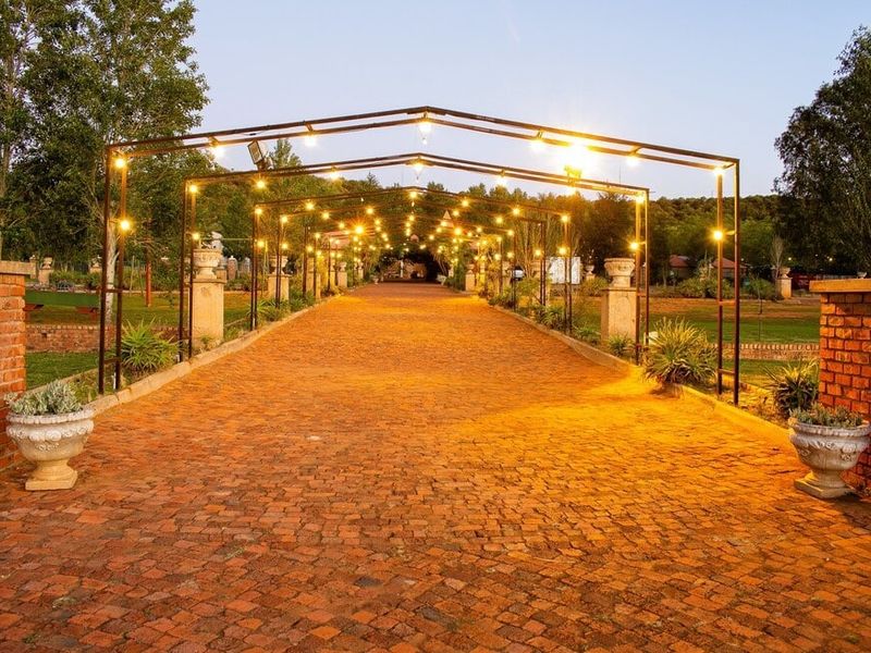 A beautifully landscaped garden pathway featuring a metal pergola structure with ambient string lighting, flanked by brick pillars and stone planters.