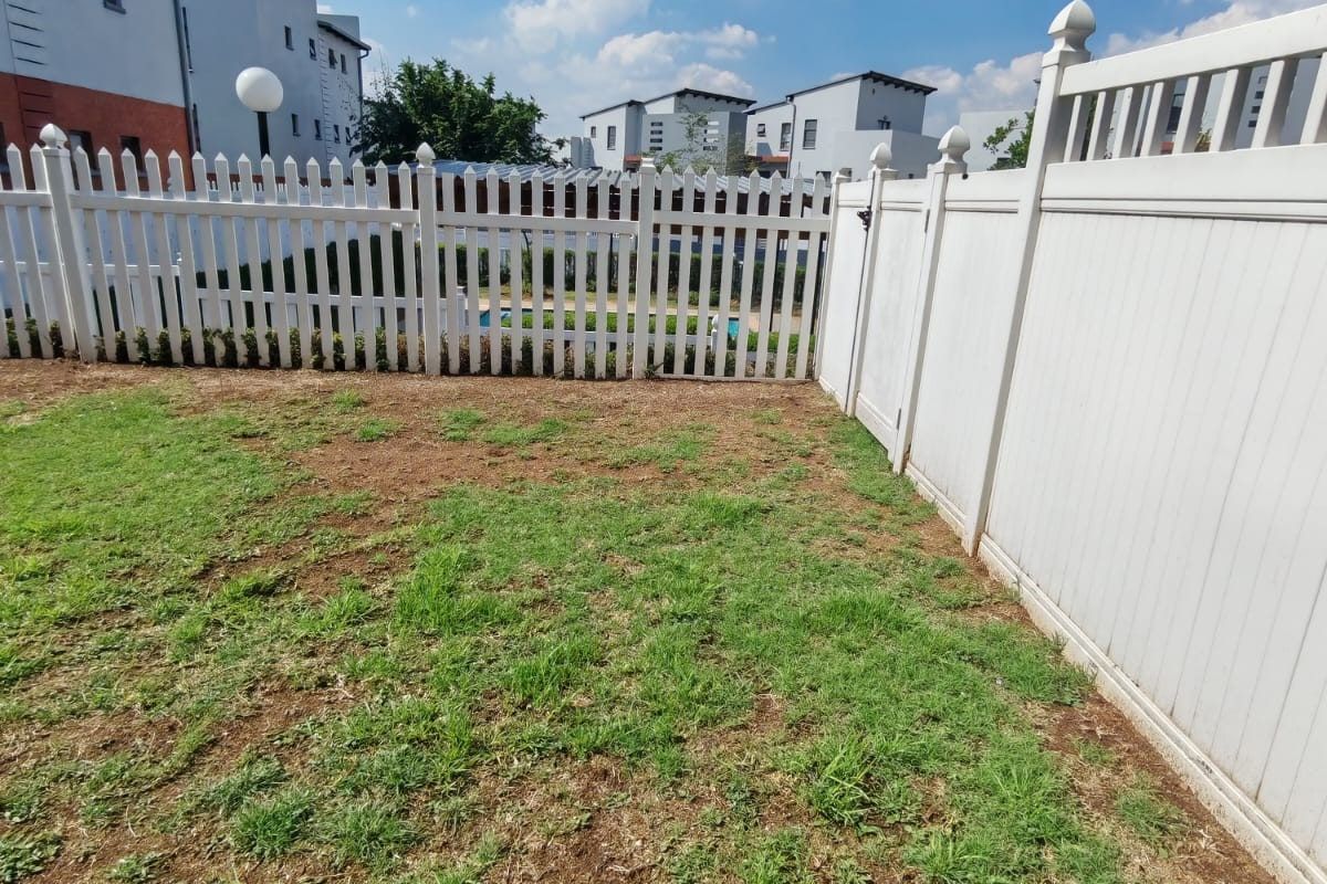 A private rear garden area enclosed by white vinyl picket fencing, featuring a lawn with some patchy areas.
