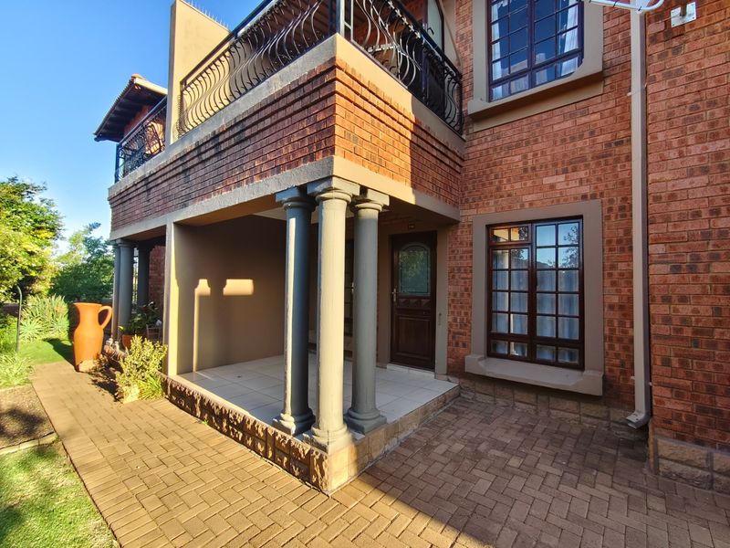 A well-maintained brick apartment exterior featuring a covered patio with decorative columns, a paved walkway, and a balcony with wrought iron railings.