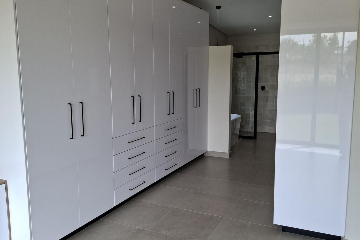 Spacious bedroom featuring extensive modern white built-in cupboards, neutral tiled flooring, and recessed downlighting, with an open-plan transition to an en-suite bathroom area.