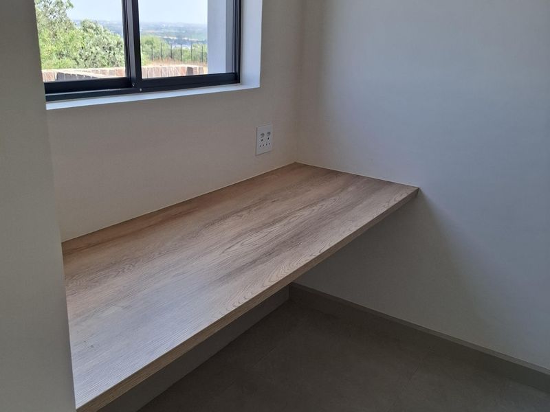 A clean, modern home office space featuring a built-in wooden desk, tiled flooring, and a large window with natural light.
