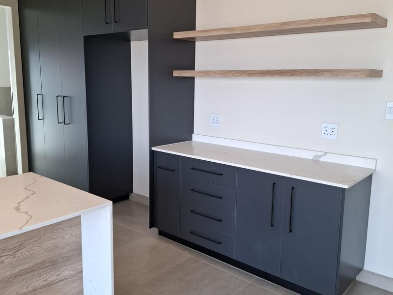 Modern kitchen featuring dark grey cabinetry, stone countertops, floating timber shelves, and a matching kitchen island with tiled flooring.