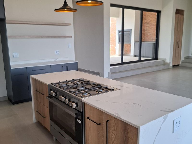 Modern open-plan kitchen featuring a large island with a gas hob, stone countertops, wood-finish cabinetry, and pendant lighting, leading to an outdoor patio area.
