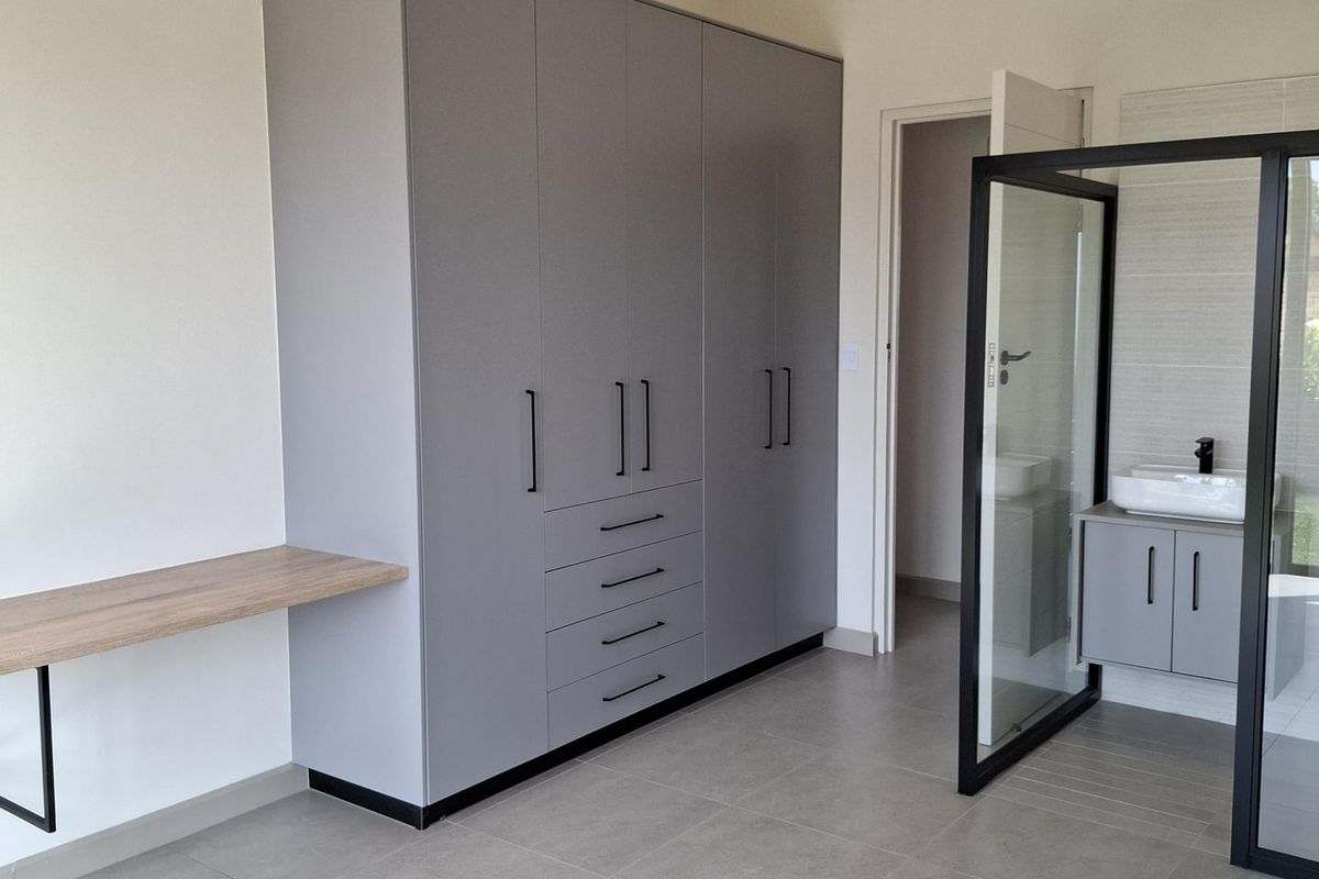 Modern bedroom featuring built-in grey cabinetry, a floating desk, and an en-suite bathroom with a glass partition and vanity.