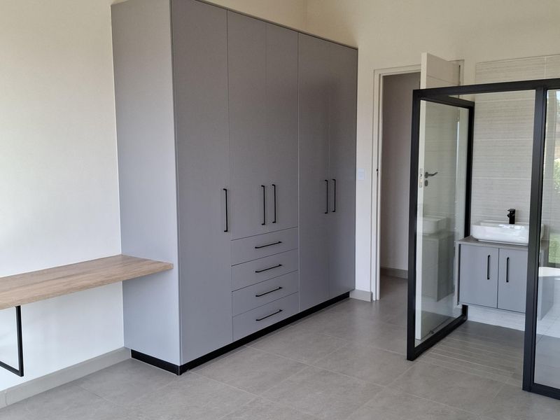 Modern bedroom featuring built-in grey cabinetry, a floating desk, and an en-suite bathroom with a glass partition and vanity.