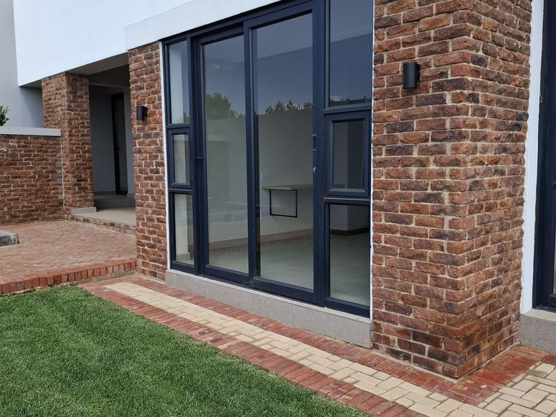 Modern exterior view featuring exposed brickwork, dark-framed sliding glass doors, and a paved walkway leading to a well-maintained lawn.