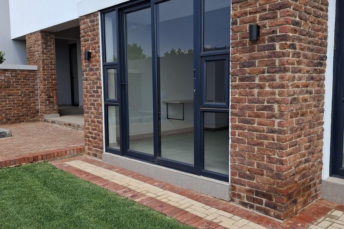 Modern exterior view featuring exposed brickwork, dark-framed sliding glass doors, and a paved walkway leading to a well-maintained lawn.