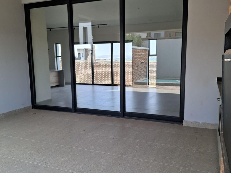 A spacious covered patio featuring large sliding glass doors, tiled flooring, and a built-in braai, offering a seamless transition to the outdoor pool area.