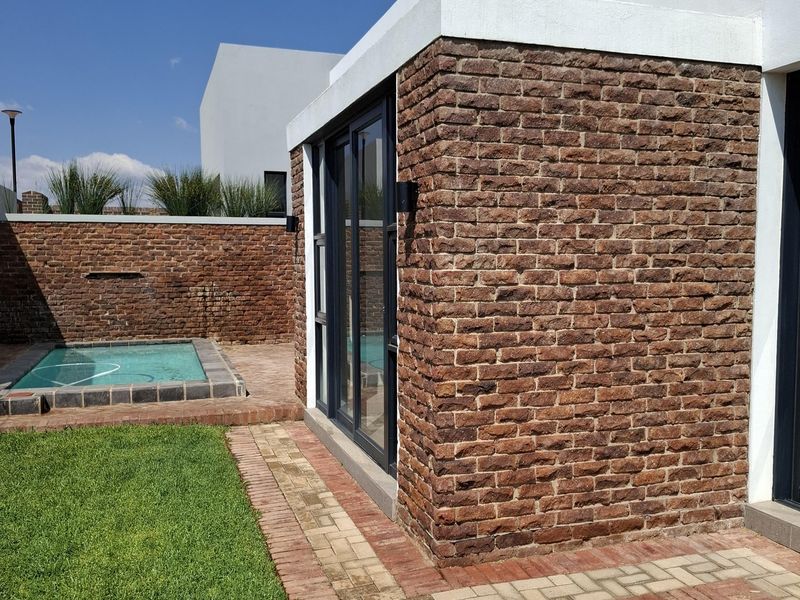 A modern rear exterior featuring an exposed brick facade, sliding glass doors, a small plunge pool, and a well-maintained lawn with brick paving.