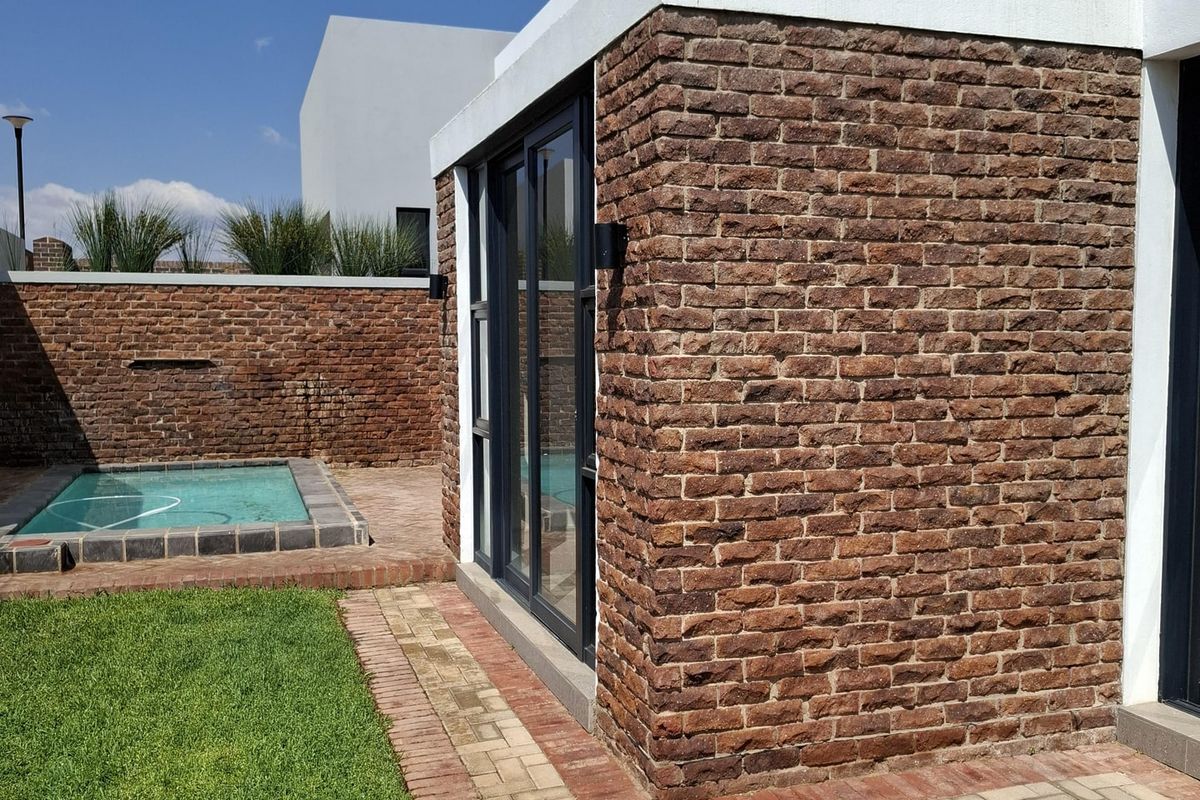 A modern rear exterior featuring an exposed brick facade, sliding glass doors, a small plunge pool, and a well-maintained lawn with brick paving.