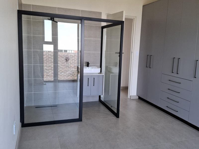 A modern, spacious bedroom featuring built-in grey cabinetry and an open-plan glass-enclosed shower and vanity area with contemporary tiled finishes.