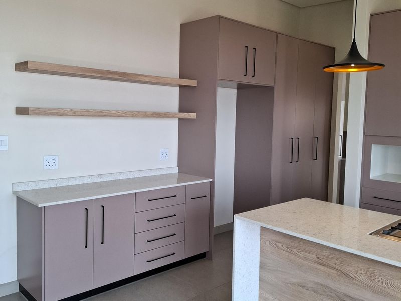 Modern kitchen featuring matte mauve cabinetry, stone countertops, a central island, floating timber shelves, and contemporary track and pendant lighting.
