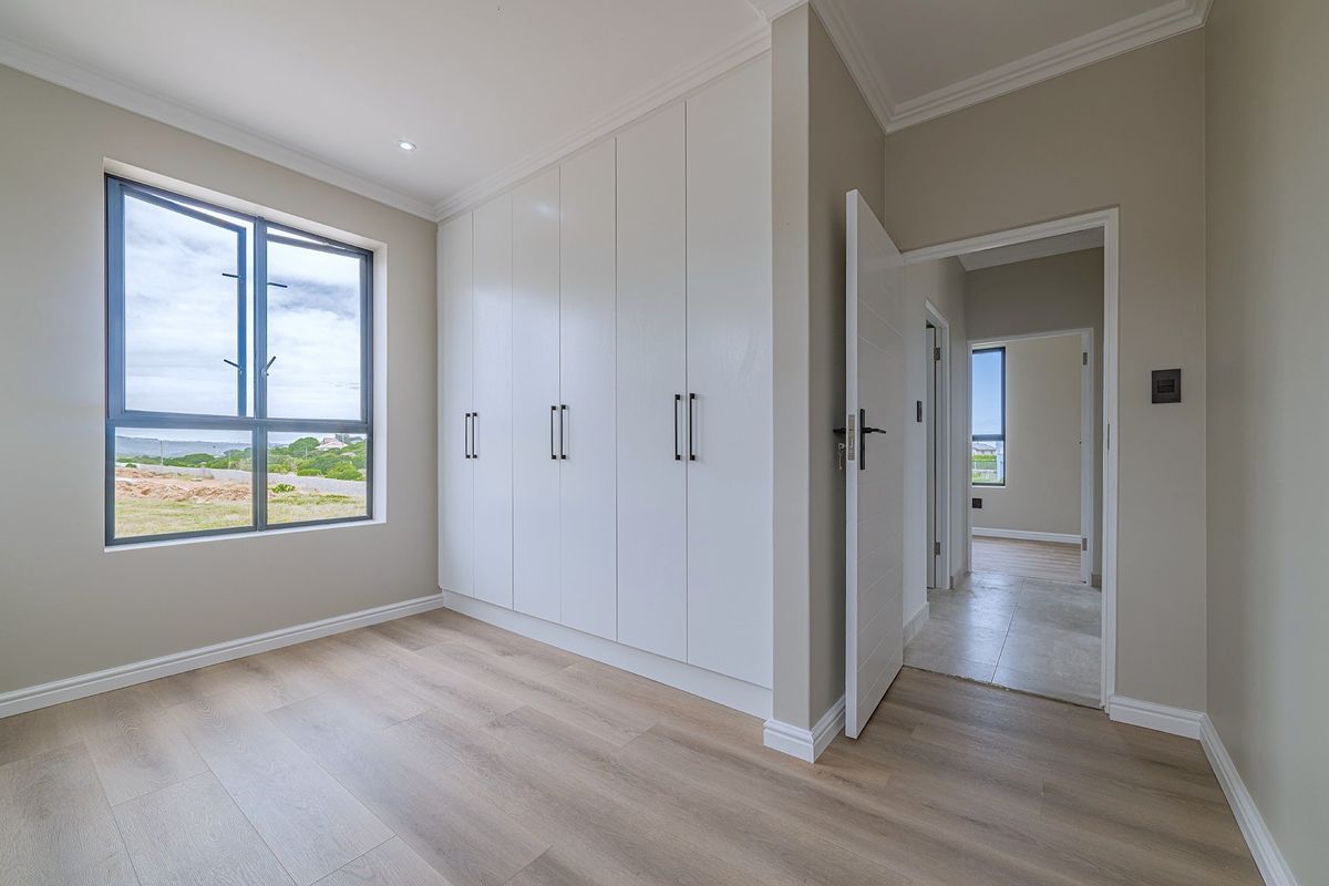 A bright, modern bedroom featuring built-in cupboards, light wood-look laminate flooring, and large aluminium windows with views of the surrounding area.