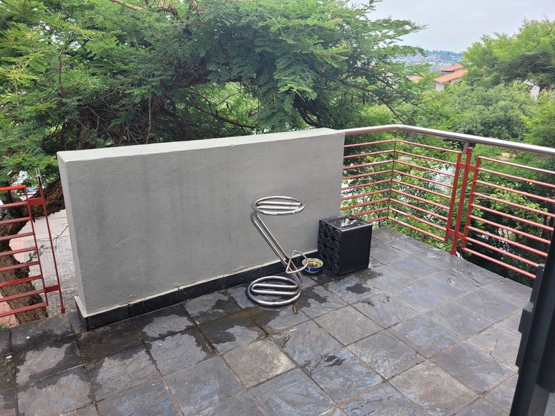 An outdoor balcony featuring slate tile flooring, a masonry privacy wall, and a metal balustrade overlooking surrounding greenery.