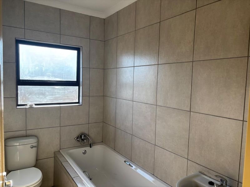 A clean, functional bathroom featuring neutral ceramic wall tiles, a built-in bathtub, a toilet, and a wall-mounted basin with natural light from the window.