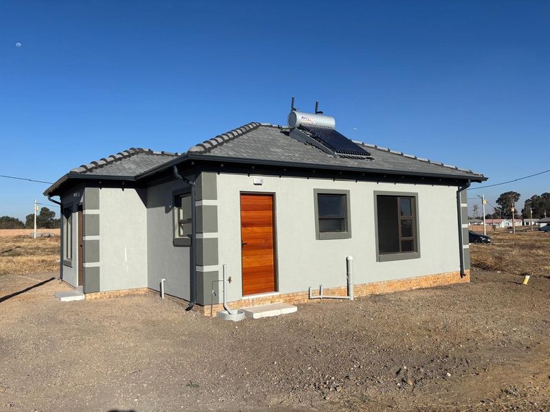 A modern single-story house exterior featuring a tiled roof, solar water heating system, timber door, and aluminium window frames set on a clear plot.