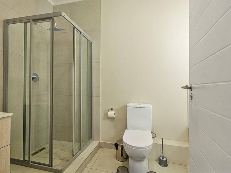 A modern bathroom featuring a glass-enclosed shower with a rain shower head, a toilet, and a vanity cabinet with neutral wall and floor tiles.