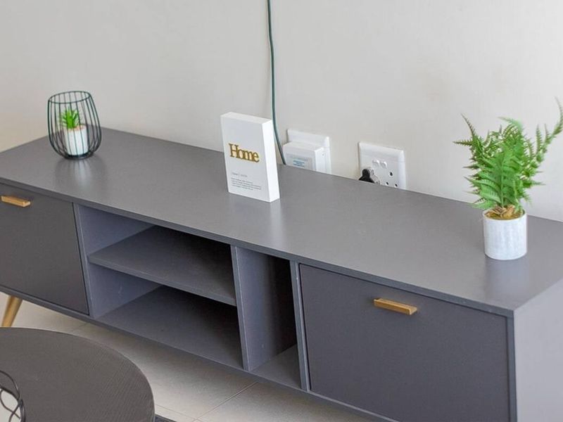 A close-up view of a modern grey media console unit positioned against a neutral wall with visible electrical power points.