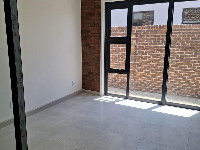 A bright, empty room featuring modern large-format floor tiles, a feature exposed brick wall, and black-framed sliding glass doors leading to an outdoor area.