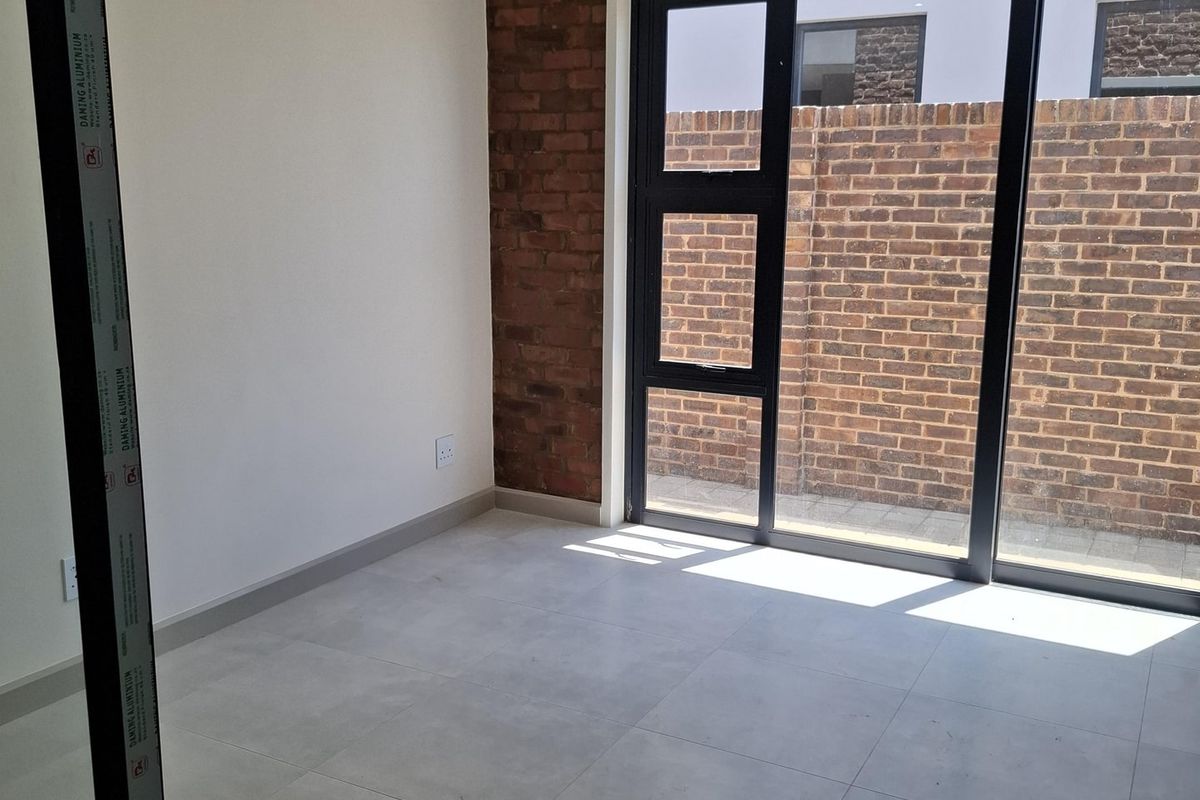 A bright, empty room featuring modern large-format floor tiles, a feature exposed brick wall, and black-framed sliding glass doors leading to an outdoor area.