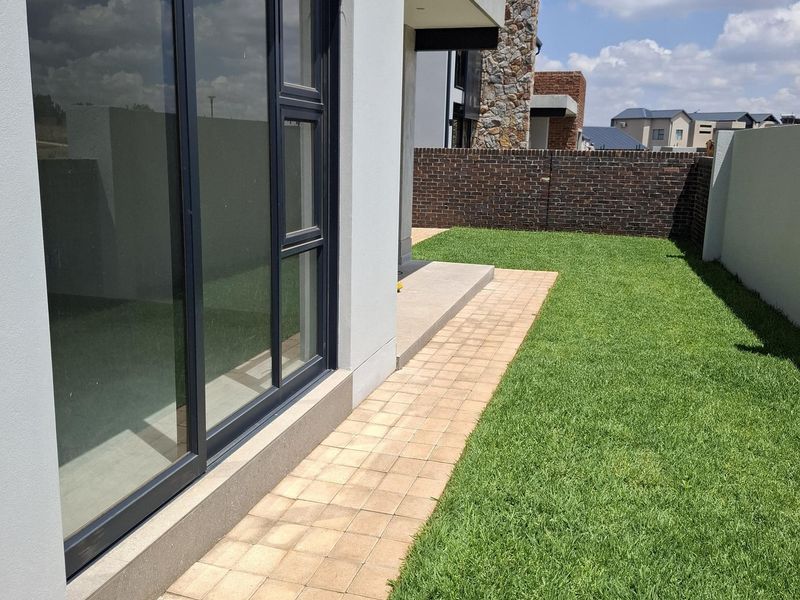 A well-maintained rear garden featuring a paved walkway, manicured lawn, and a brick boundary wall, adjacent to modern sliding glass doors.