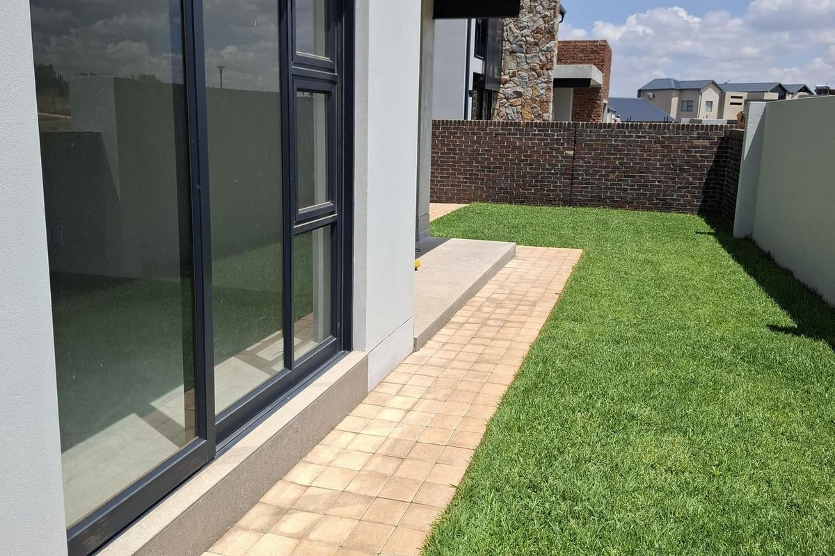 A well-maintained rear garden featuring a paved walkway, manicured lawn, and a brick boundary wall, adjacent to modern sliding glass doors.