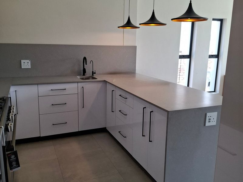 Modern kitchen featuring sleek white cabinetry, grey quartz countertops, tiled flooring, and contemporary pendant lighting in a clean, minimalist design.