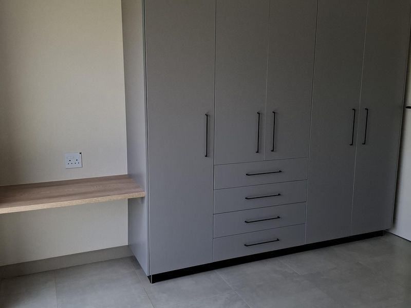 A modern bedroom featuring a large grey built-in wardrobe with integrated drawers and a matching floating desk, complemented by neutral tiled flooring.