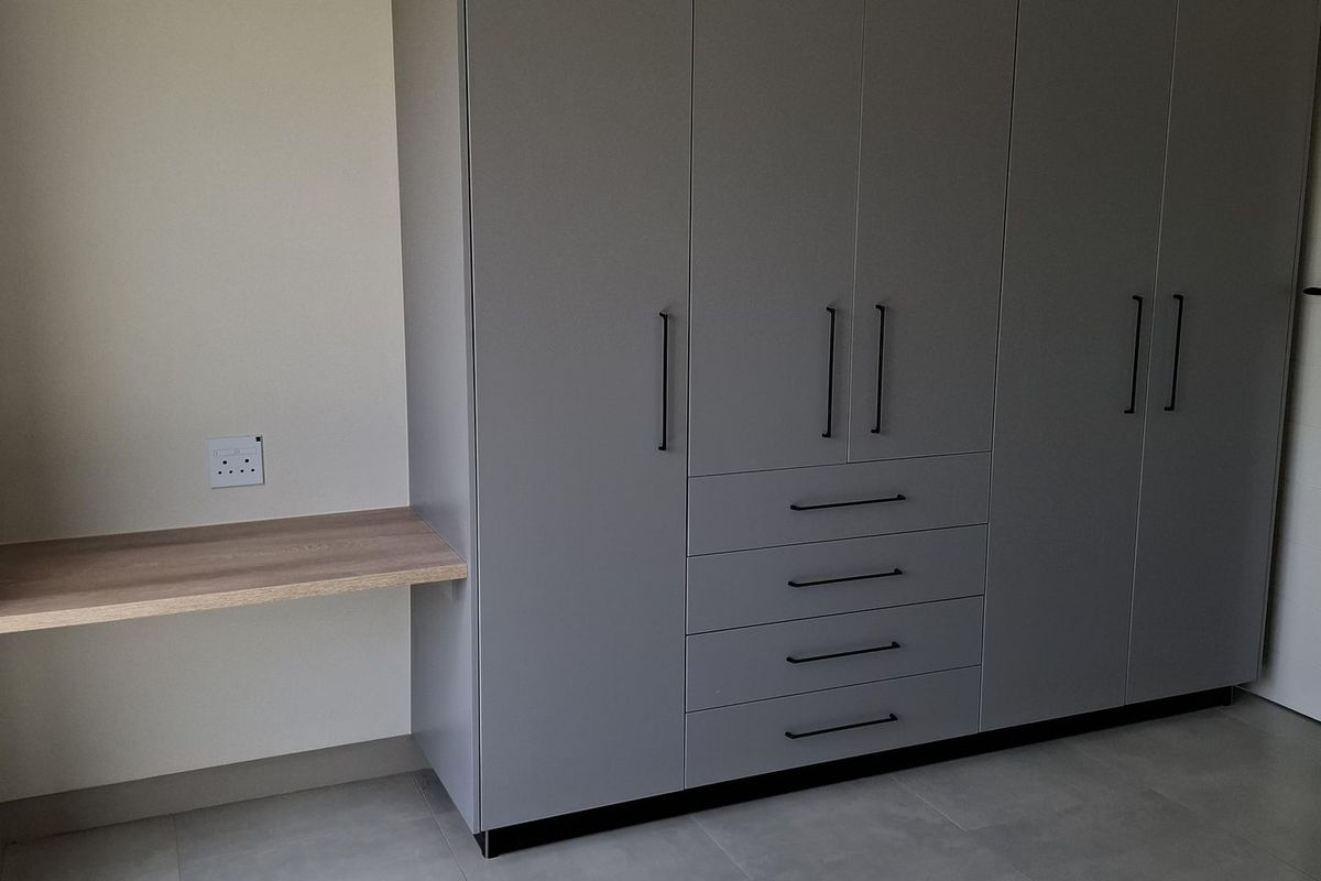 A modern bedroom featuring a large grey built-in wardrobe with integrated drawers and a matching floating desk, complemented by neutral tiled flooring.