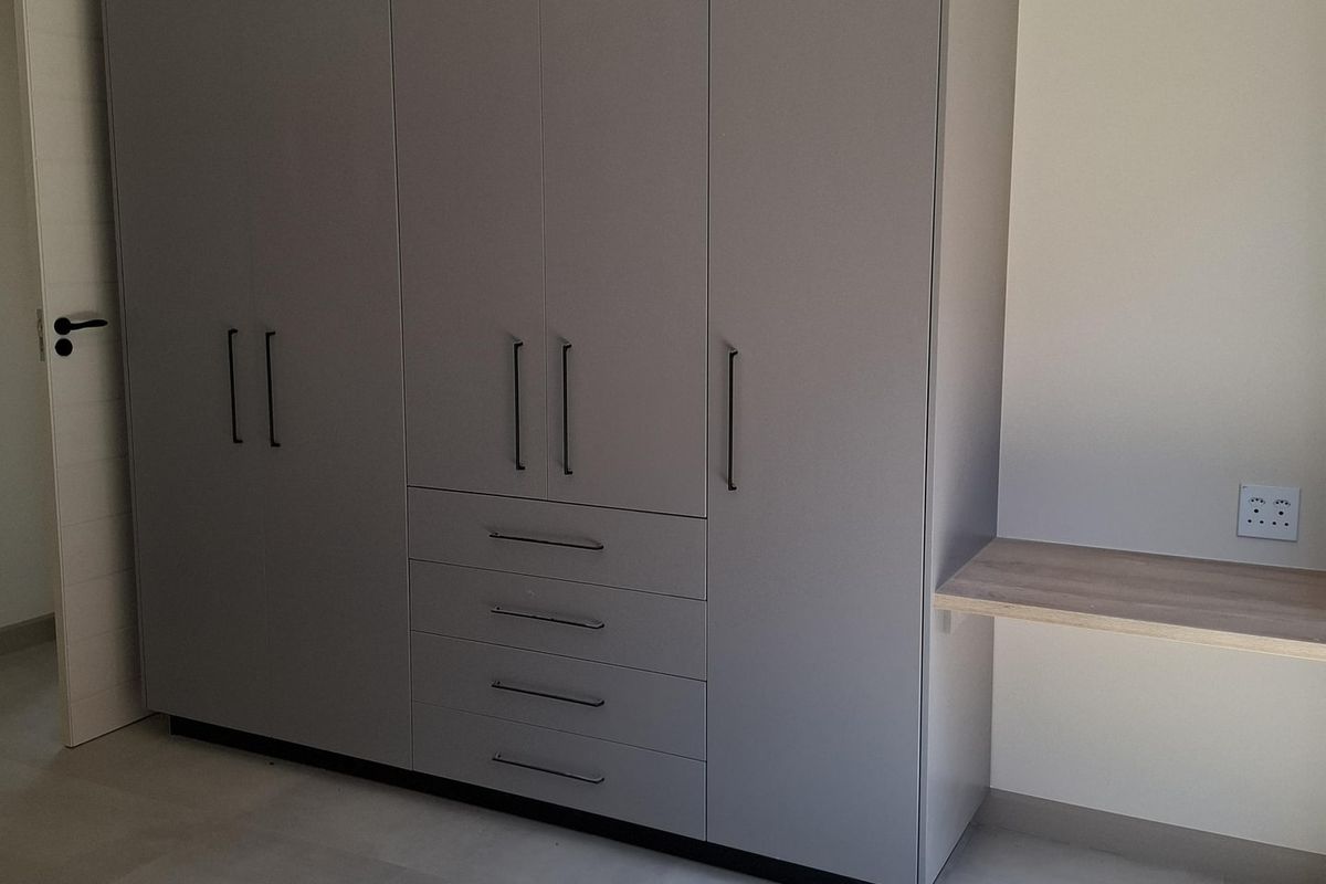 A modern bedroom featuring a large grey built-in wardrobe with integrated drawers and a floating desk shelf, finished with neutral floor tiles.
