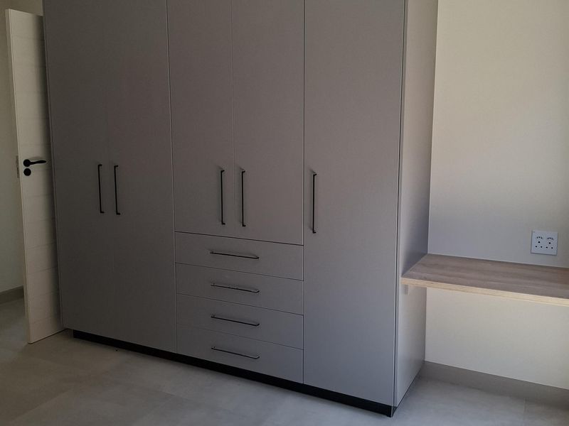 A modern bedroom featuring a large grey built-in wardrobe with integrated drawers and a floating desk shelf, finished with neutral floor tiles.