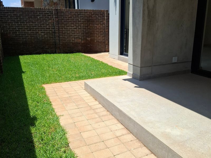 A private rear garden featuring a neat lawn, paved walkway, and a covered concrete patio area enclosed by a brick boundary wall.