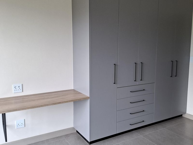 A modern bedroom featuring a large grey built-in wardrobe with integrated drawers and a matching floating desk, set against neutral walls and tiled flooring.