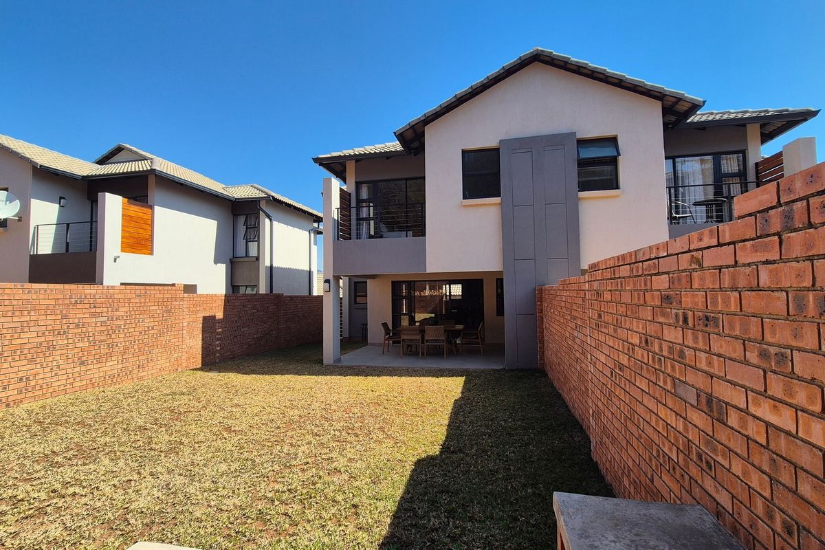 Rear view of a modern townhouse featuring a private garden, brick boundary walls, a covered patio, and an upper-level balcony.
