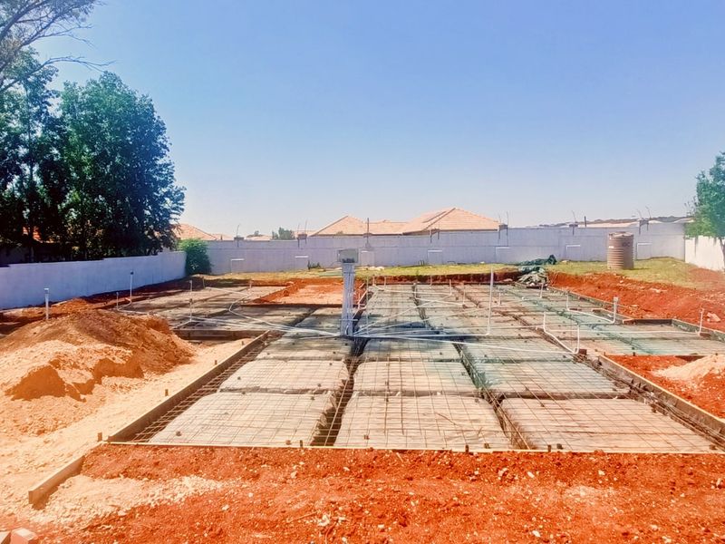 A construction site showing a prepared concrete foundation slab with steel reinforcement and plumbing conduits installed, surrounded by a perimeter boundary wall.
