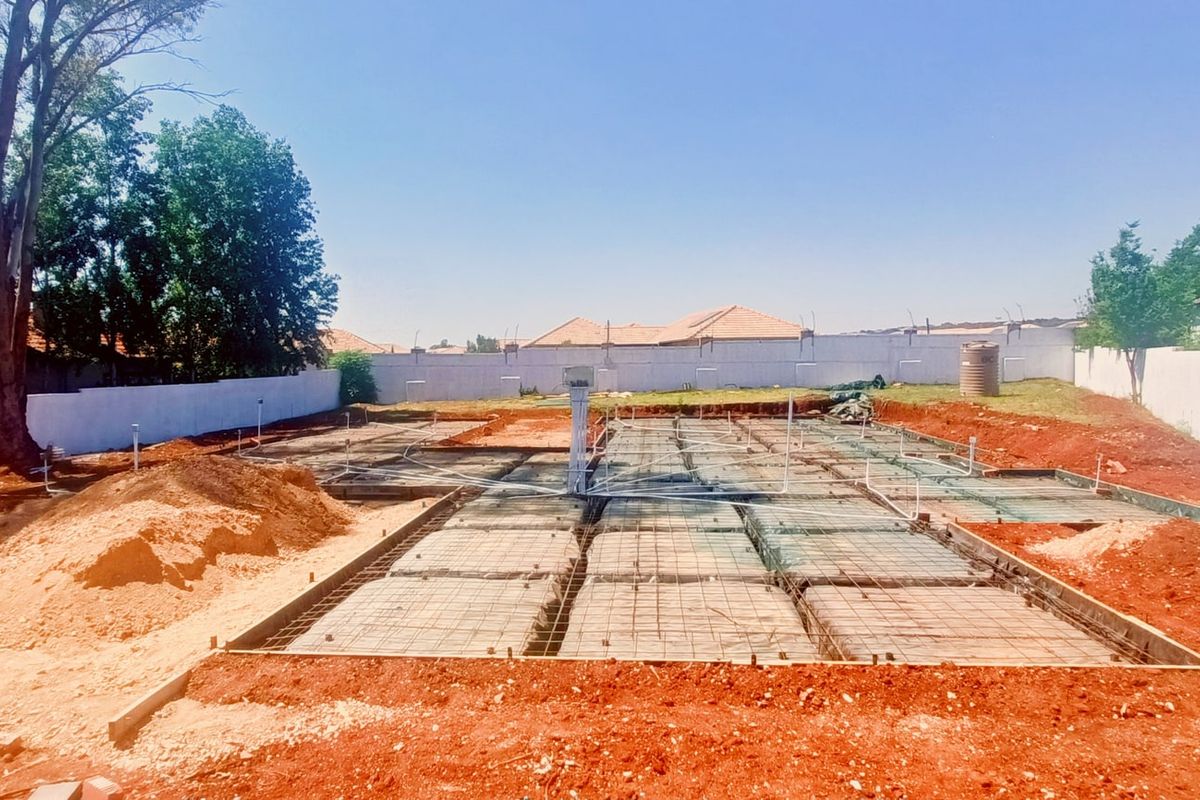 A construction site showing a prepared concrete foundation slab with steel reinforcement and plumbing conduits installed, surrounded by a perimeter boundary wall.