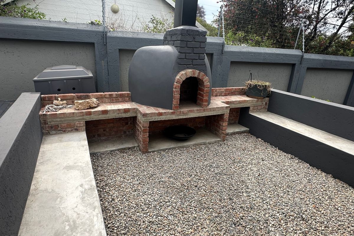 Outdoor entertainment area featuring a built-in brick pizza oven, concrete bench seating, and a gravel courtyard enclosed by a perimeter boundary wall.