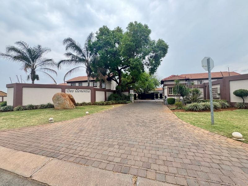 The entrance to the Deo Clementia townhouse complex features a paved driveway, secure perimeter wall with electric fencing, and well-maintained landscaped gardens.