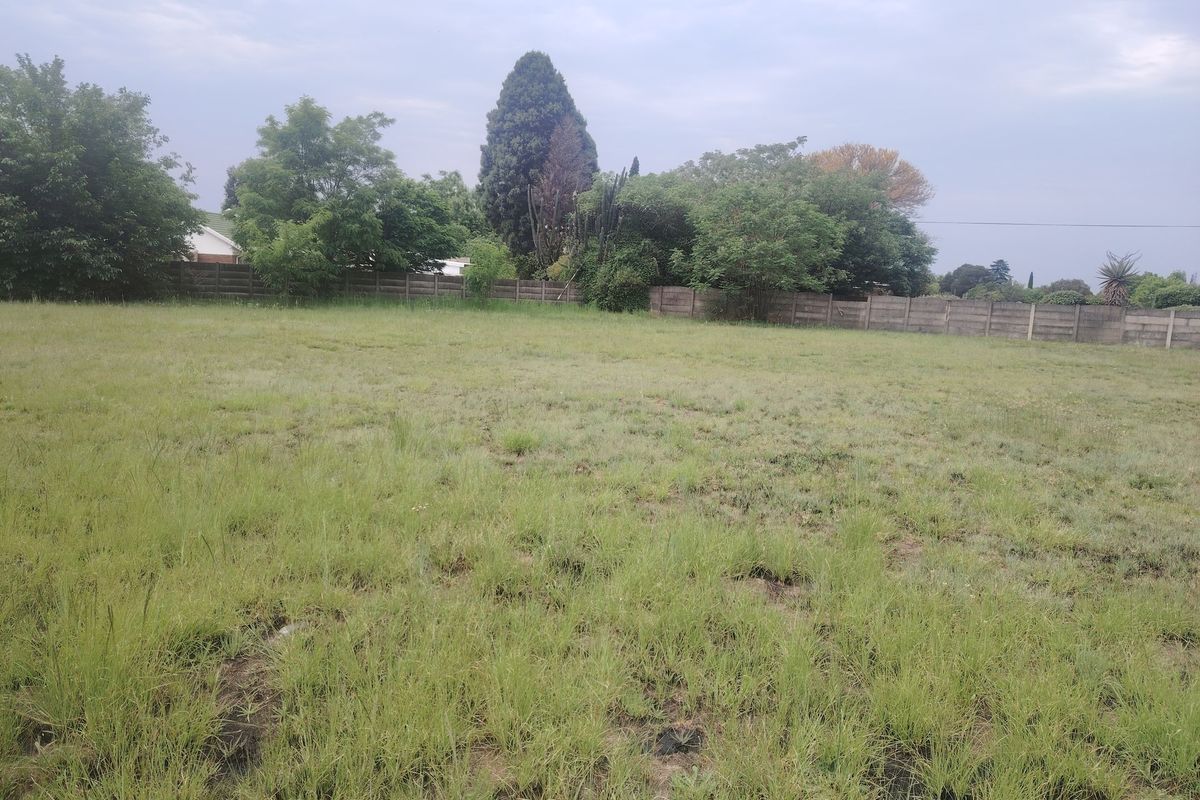 A vacant residential plot featuring a grass lawn, mature trees, and a precast concrete boundary wall at the rear.