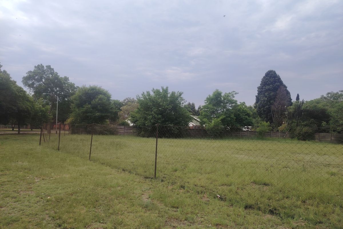 A view of a vacant residential plot featuring a grass lawn, mature trees, and perimeter wire mesh fencing.
