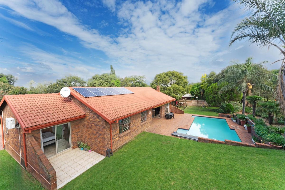 Rear view of a brick house featuring a swimming pool, paved patio area, and roof-mounted solar panels set within a landscaped garden.
