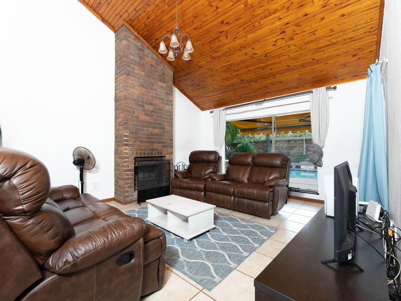 Spacious living room featuring a prominent brick fireplace, vaulted timber ceiling, and tiled flooring with direct access to the outdoor pool area.